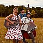 Die Schlagergang steht lachend auf einer Wiese: eine Frau mit Gitarre im Kirschkleid, ein Mann mit Akkordeon und ein Kontrabassist mit Hut, im Hintergrund Wald und Himmel.