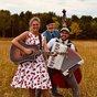 Die Schlagergang steht lachend auf einer Wiese: eine Frau mit Gitarre im Kirschkleid, ein Mann mit Akkordeon und ein Kontrabassist mit Hut, im Hintergrund Wald und Himmel.