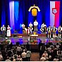 Menschen in traditioneller Tracht stehen und sitzen auf einer Bühne. Im Hintergrund hängen blau-weiße und rot-weiße Fahnen. Ein Publikum in Tracht schaut zur Bühne. Es handelt sich um eine Volksmusikveranstaltung.