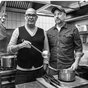 Das schwarz-weiß gefärbte Foto zeigt Hannes Ringlstetter und Stephan Zinner in einer Großküche mit einer Kelle und einem Topf in der Hand.