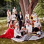 Max Raabe in Smoking mit dem Palast Orchester in eleganter Kleidung, bei einem Picknick unter einem großen Baum, umgeben von Instrumenten wie Tuba, Trompete und Geige.