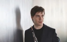 Person steht vor hellem Hintergrund und hält ein Holzblasinstrument in der Hand.